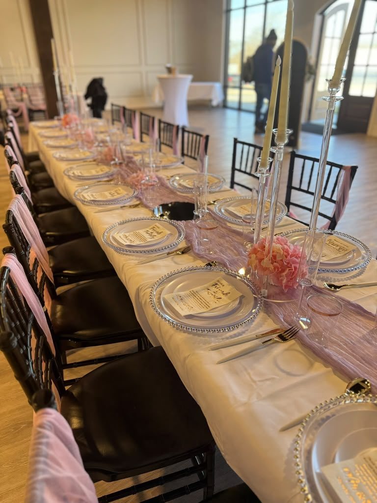 Long banquet table with white linens, lavender runner, gold flatware, taper candles, pink hydrangeas, black Chiavari chairs