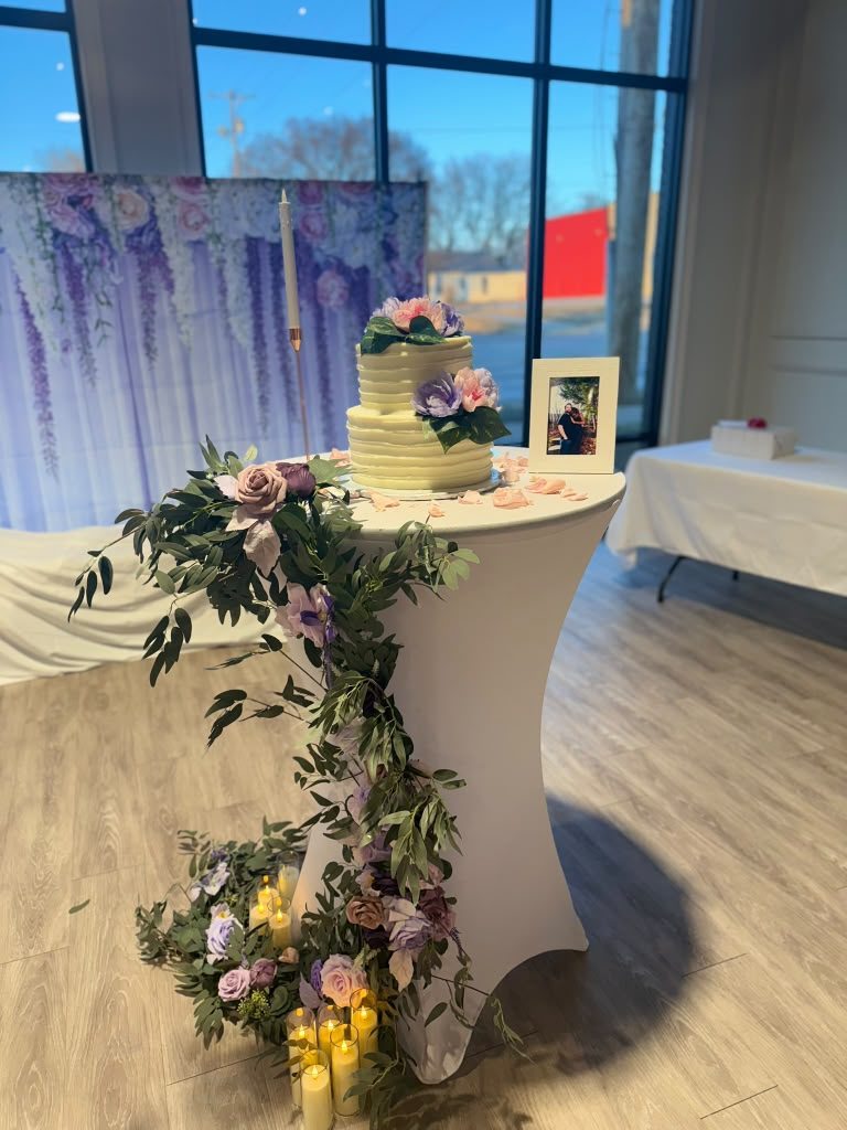 Two-tier ivory cake with purple and pink florals on a cocktail table, cascading eucalyptus garland and candle cylinders