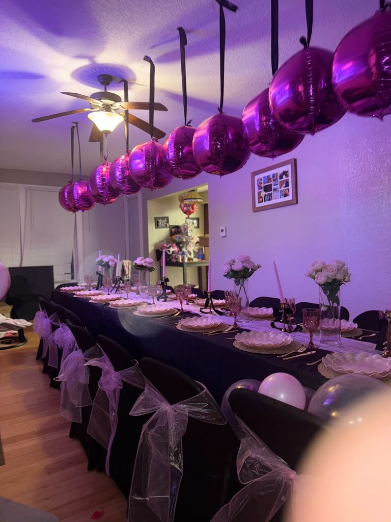 Black-linen dinner table with gold flatware, rose centerpieces, magenta foil balloons overhead